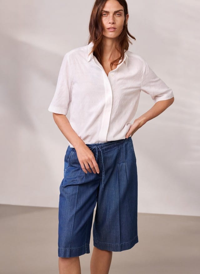 Eine Person trägt ein weißes Kurzarmhemd und knielange blaue Jeansshorts und steht vor einer neutralen Wand. Eine Person trägt ein weißes Kurzarmhemd und knielange blaue Jeansshorts und steht vor einer neutralen Wand.