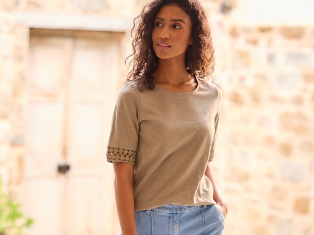 Frau in einem beigen T-Shirt steht vor einer Steinmauer und schaut zur Seite.