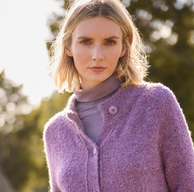Das Bild zeigt eine Frau, die eine lilafarbene Strickjacke mit Knöpfen, einen passenden Rollkragenpullover und eine weiße Hose trägt und sich im Freien aufhält.