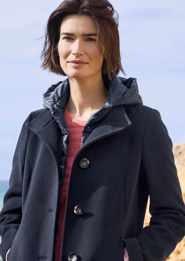 Eine Frau in einer dunkelblauen Wolljacke mit Kapuze über einem lachsfarbenen Pullover steht vor Felsen am Meer und schaut in die Kamera.