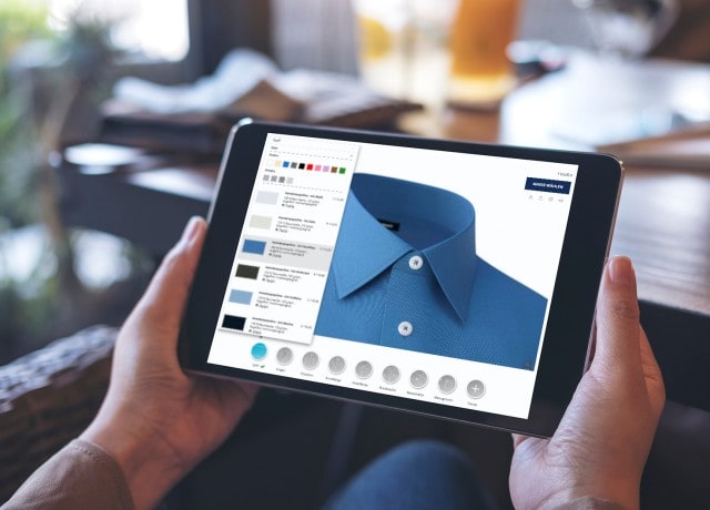 Auf dem Bild hält eine Person ein Tablet in den Händen, auf dessen Bildschirm ein Online-Konfigurator für Hemden mit verschiedenen Auswahlmöglichkeiten für Stoffe, Farben und Kragenformen angezeigt wird.