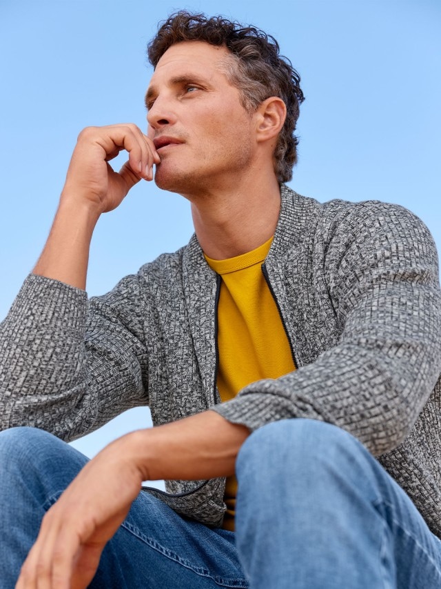 Ein Mann sitzend, in Jeans, gelben Shirt und Strickjacke.