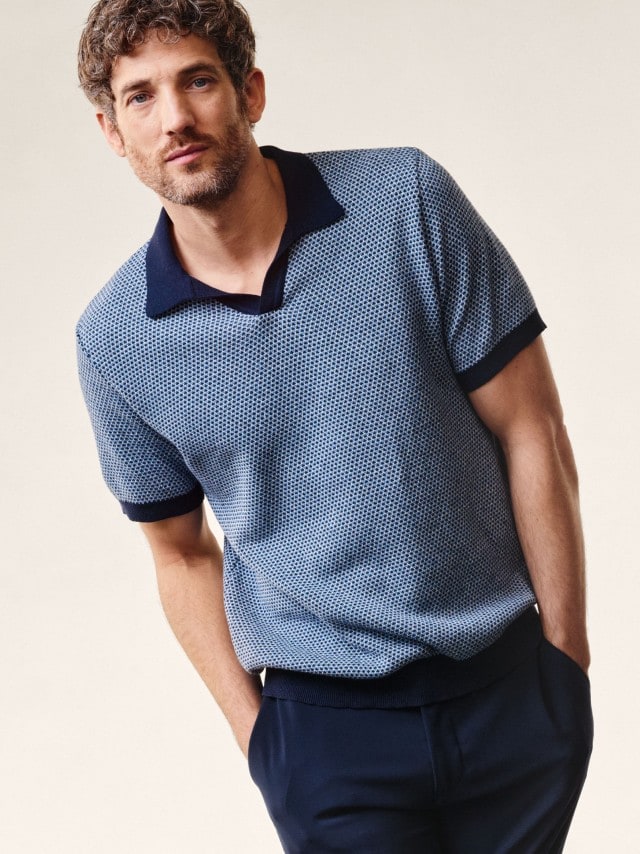 Ein Mann trägt ein hellblaues Strickpolo und eine dunkelblaue Hose.