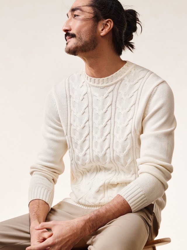 Mann sitzend vor neutralem Hintergrund trägt den Zopfpullover Woolcot dazu die Macht-Alles-Mit Hose 2.0 in Beige.