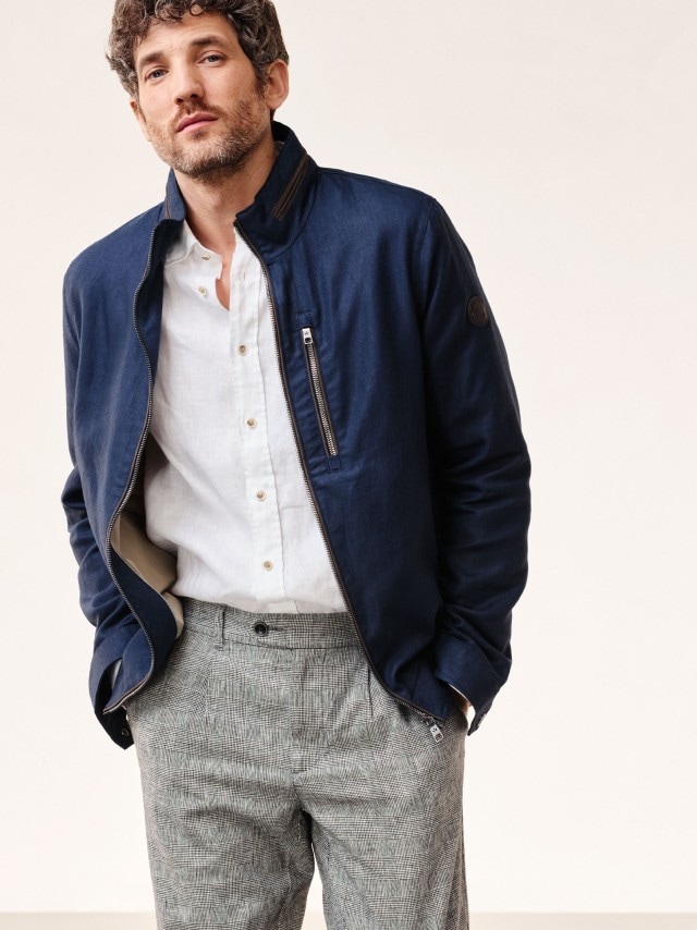 Ein Mann trägt einen marinefarbenden Blouson mit weißem Hemd und gemusterter Hose.
