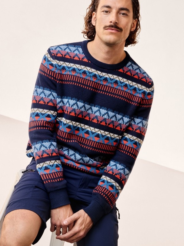 Ein Mann sitzend, trägt einen gemusterten Jacquard-Pullover, dazu eine Bermudas in marine. 