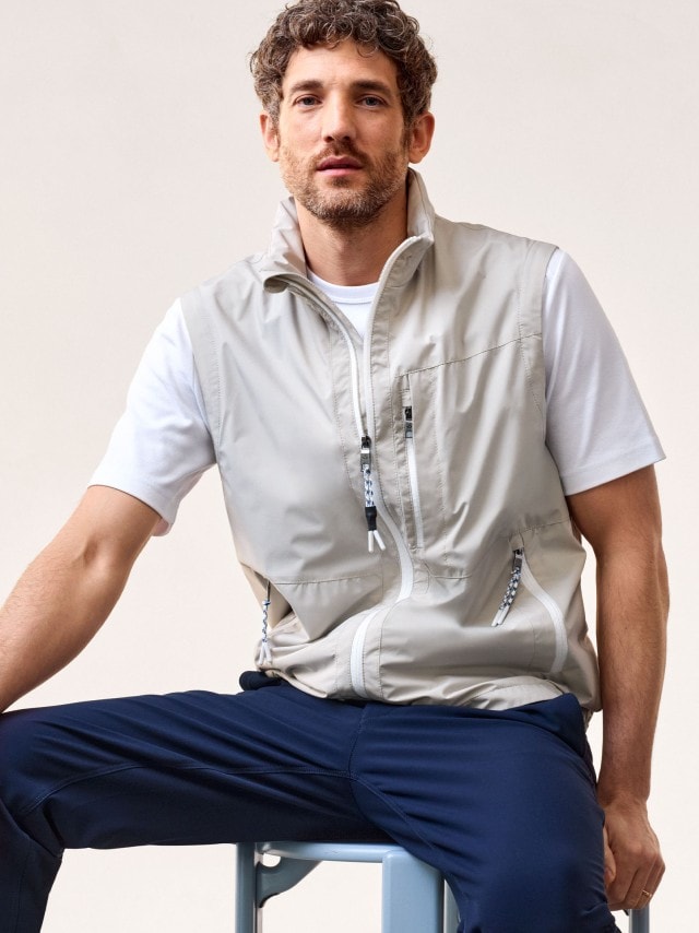 Ein Mann sitzt auf einen Hocker und trägt eine Weste in beige, darunter ein weißes T-Shirt. 