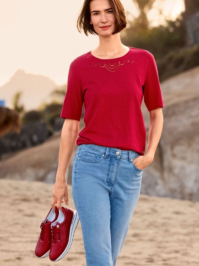 Frau spaziert am Strand und trägt ein Baumwollshirt Lochstickerei in Rot, dazu eine blaue Jeans und hält ihre Schuhe in der Hand