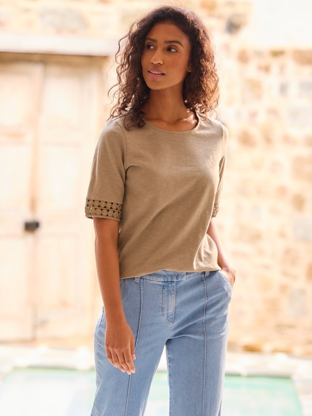 Eine Frau steht draußen und trägt ein beigefarbenes T-Shirt mit dekorativen Spitzenärmeln. Dazu kombiniert sie eine hellblaue Jeans mit sichtbaren Nähten.