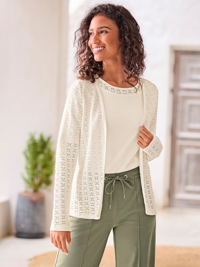 Eine Fra trägt einen cremefarbenen, durchbrochenen Cardigan über einem passenden, unifarbenen Top. Dazu kombiniert sie eine olivgrüne Hose mit einem Kordelzug an der Taille.