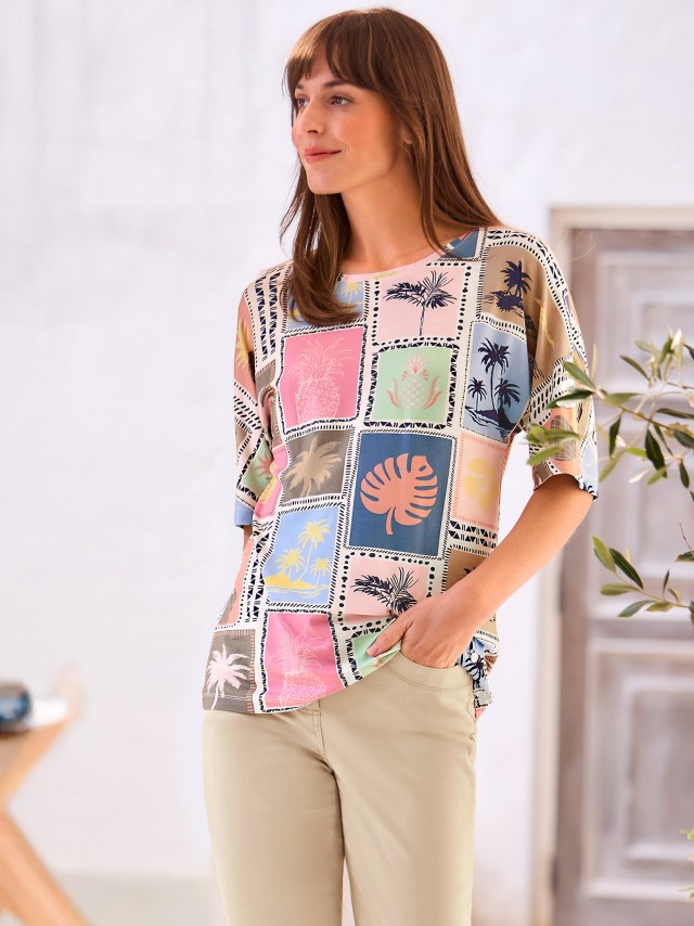 Eine Frau trägt ein bunt bedrucktes T-Shirt mit tropischen Motiven in sommerlichen pastellfarben. Sie kombiniert es mit beigen Hosen.