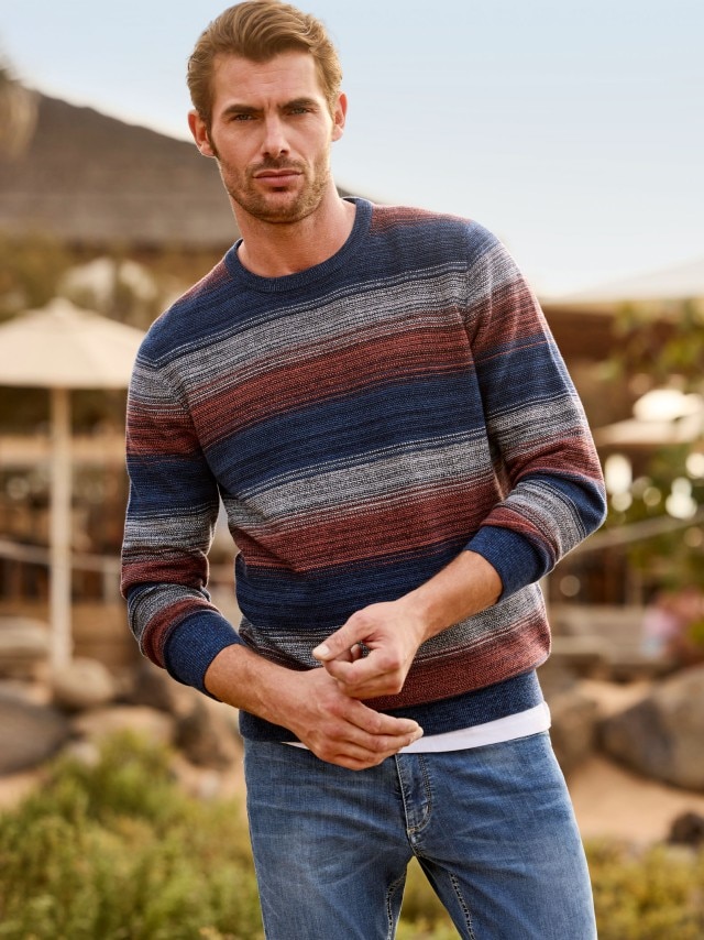 Mann steht am felsigen Strand und trägt einen Struktur Pullover mit blauer Jeans