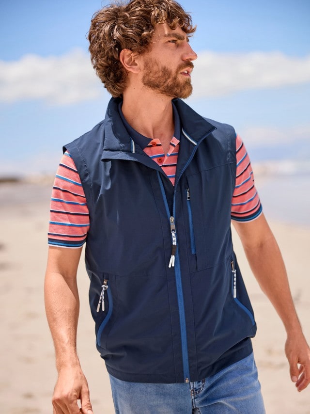 Ein Mann trägt eine sportliche, dunkelblaue Weste über einem gestreiften Poloshirt in Blau und Korallrot. Dazu kombiniert er eine lässige Jeans.