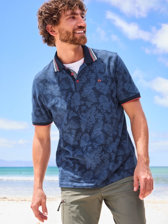 Ein Mann trägt ein dunkelblaues Poloshirt mit dezentem floralen Muster und roten Akzenten am Kragen, kombiniert mit einer khakifarbenen Cargohose. Der Look ist lässig, modern und ideal für einen entspannten Tag am Strand.