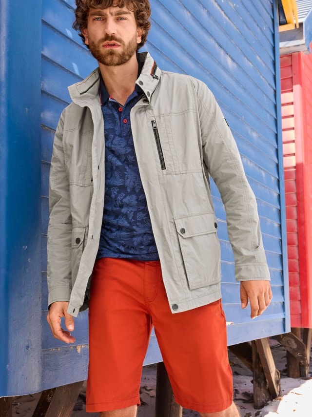 Ein Mann trägt eine hellgraue Sommerjacke über einem dunkelblauen Poloshirt mit floralem Muster und kombiniert dazu leuchtend orange Shorts. Der Look ist modern, frisch und perfekt für einen entspannten Tag am Strand.