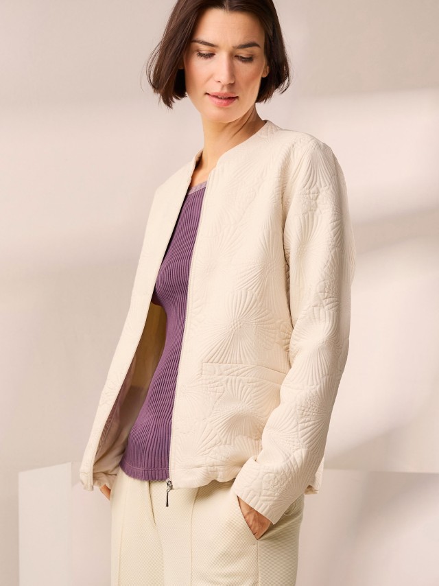 Eine Person trägt eine cremefarbene, strukturierte Jacke mit floralem Muster über einem violetten Oberteil und heller Hose.