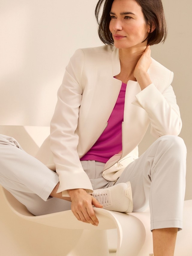 Eine Person sitzt entspannt auf einem hellen Stuhl und trägt einen weißen Blazer über einem pinkfarbenen Oberteil, dazu helle Hose und weiße Sneaker.