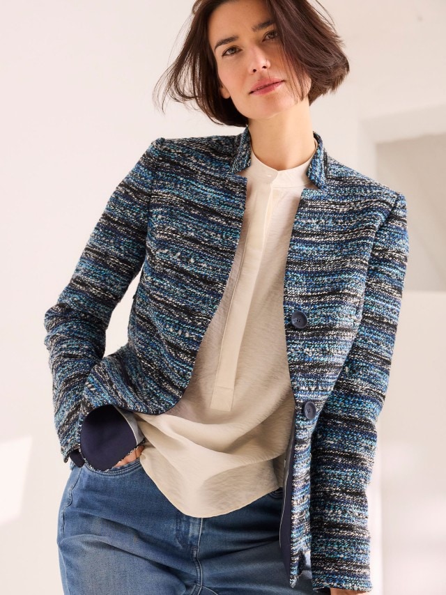 Eine Person trägt eine blau gemusterte, strukturierte Jacke über einer hellen Bluse und kombiniert sie mit einer blauen Jeans.