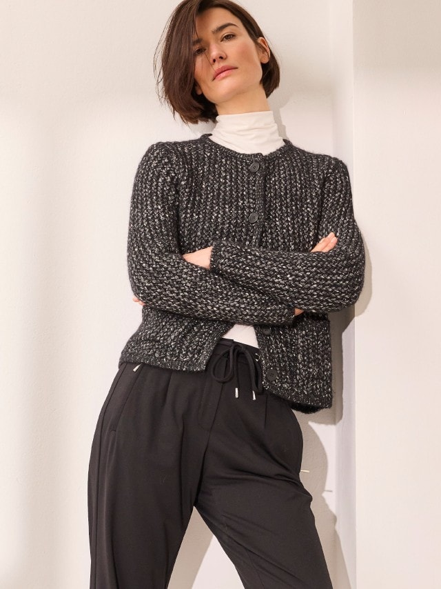 Eine Person trägt einen weißem Rollkragenpullover unter einer grob gestrickten, schwarz-grauen Strickjacke und kombiniert dazu eine schwarze Hose mit Kordelzug.