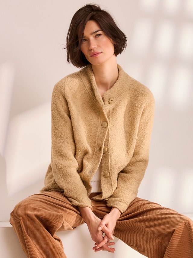 Eine Person sitzt entspannt und trägt eine beige, flauschige Strickjacke mit großen Knöpfen, kombiniert mit einer gerippten, braunen Cordhose.