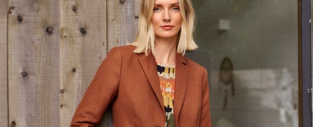 Eine Frau steht vor einer Holzwand und trägt einen braunen Blazer. Darunter trägt sie ein bunt gemustertes Oberteil. Der Hintergrund ist ein Fenster mit Blick in einen Innenraum.