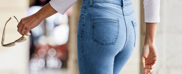Eine Person trägt eine hellblaue Jeans und hält eine Sonnenbrille in der Hand. Sie hat ein weißes Oberteil an. Der Fokus liegt auf der Rückansicht der Jeans.