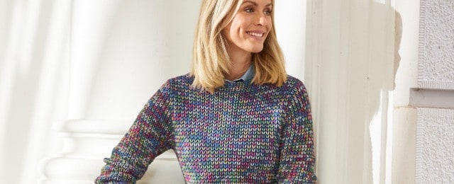 Eine Frau trägt einen bunt gestrickten Pullover mit einem blauen Hemd darunter. Sie lehnt an einem weißen Säulenhintergrund und lächelt.