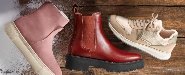 Drei verschiedene Schuhe auf einem Holzboden: ein rosa Stiefel mit weißer Sohle, ein roter Lederstiefel mit schwarzer Plateau-Sohle und ein beigefarbener Sneaker mit flauschigem Innenfutter.
