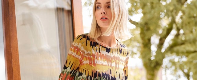Eine Frau trägt einen farbenfrohen Pullover mit abstraktem Muster. Sie steht neben einer Glastür mit Blick auf die Natur im Hintergrund.