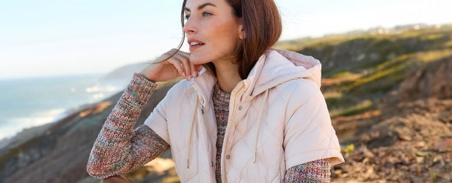 Eine Frau sitzt in einer Landschaft mit Küstenblick und trägt eine hellrosa Steppweste mit Kapuze über einem bunten Strickpullover. Sie blickt nachdenklich in die Ferne.