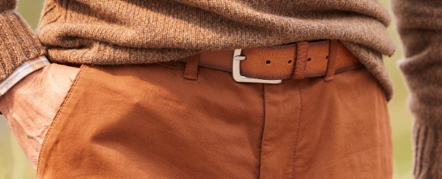 Ein Detailfoto eines Mannes in einem braunen Strickpullover, der über einem karierten Hemd getragen wird. Er trägt eine hellbraune Hose mit einem passenden Ledergürtel. Seine Hand steckt locker in der Hosentasche.