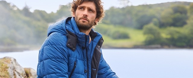 Ein Mann sitzt draußen in einer Naturkulisse und trägt eine blaue Daunenjacke mit Kapuze. Im Hintergrund sind verschwommene Bäume und ein See zu sehen.