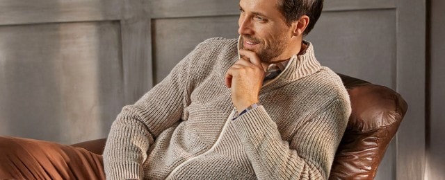 Ein Mann sitzt entspannt in einem braunen Ledersessel und trägt einen beigen Strickpullover mit Reißverschluss. Er schaut zur Seite und wirkt nachdenklich. Der Hintergrund besteht aus einer grauen Holzwand.
