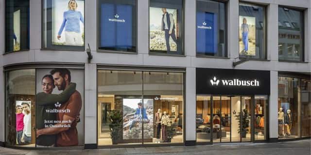 Die Aufnahme zeigt die Außenansicht eines Walbusch-Modegeschäfts mit großen Schaufenstern und Werbeplakaten, durch die man den Verkaufsraum sehen kann.