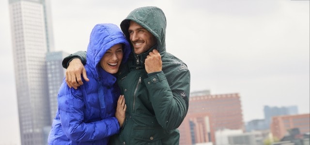 Lachendes Paar in wetterfesten Jacken mit Kapuzen umarmt sich vor städtischer Kulisse.