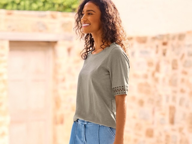 Eine junge Frau mit lockigen Haaren steht lächelnd draußen vor einer hellen Steinmauer und trägt ein Shirt mit Spitzenmuster an den Ärmeln in Khaki sowie eine blaue Hose.