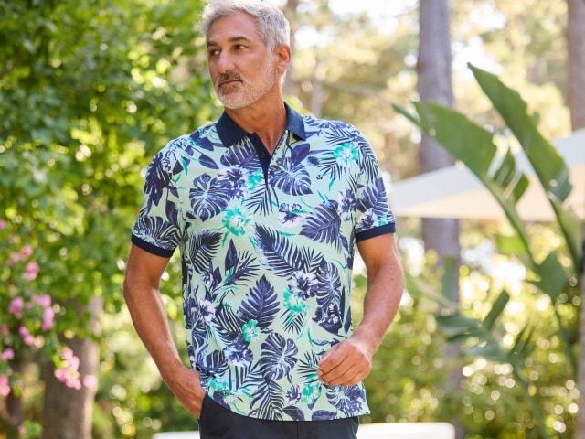 Ein Mann trägt ein kurzärmliges, gemustertes Polo-Shirt mit tropischen Blumen und Blättern.