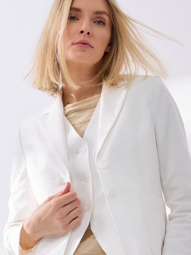 Eine blonde Frau trägt einen weißen Seersucker Blazer, mit passender Weste und Hose, dazu ein Baumwollshirt mit Streifenstruktur in Beige. 