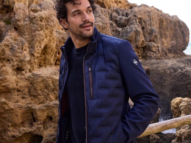 Ein Mann vor einer Felswand trägt eine steppte Jacke in Marine, darunter einen passenden Pullover.