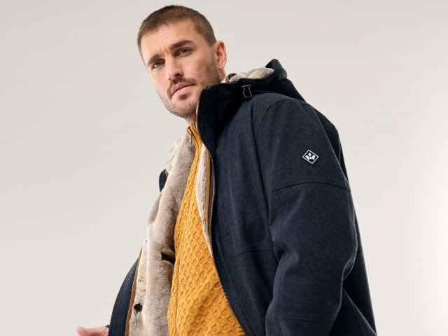 Ein Mann trägt eine dunkelgraue Jacke mit Kapuze und hellem Innenfutter über einem senfgelben Strickpullover.