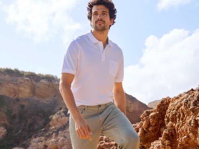 Ein Mann trägt ein weißes Poloshirt mit Brusttasche und eine helle Hose in einer Felslandschaft.