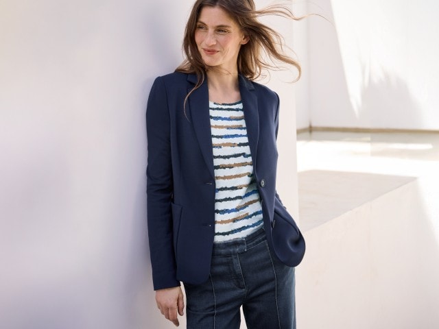 Eine Frau trägt einen dunkelblauen Blazer über einem gestreiften Oberteil und einer dunklen Jeans.