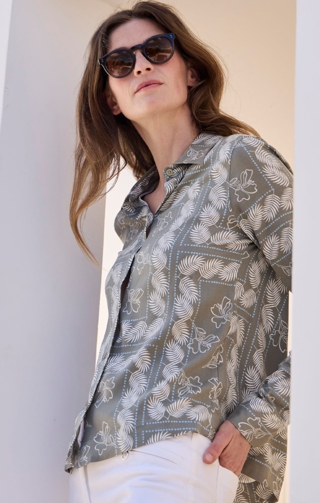 Eine Frau mit Sonnenbrille, die eine grau-grüne Bluse mit weißem floralem Muster trägt, dazu eine weiße Hose.