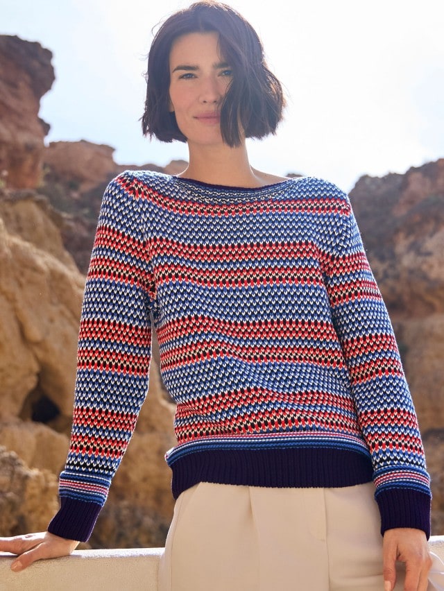 Eine Frau steht an einer Felsküste und trägt einen bunt-gemusterten Strickpullover, dazu eine Hose in Beige.