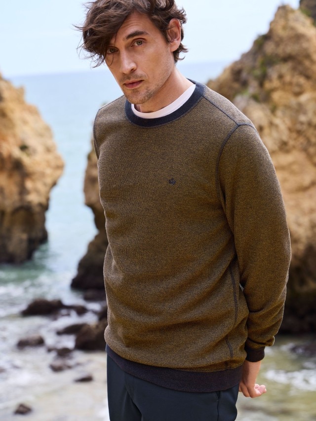 Ein Mann steht an einer Felsküste und trägt einen Struktur-Pullover, dazu eine Hose in Marine.