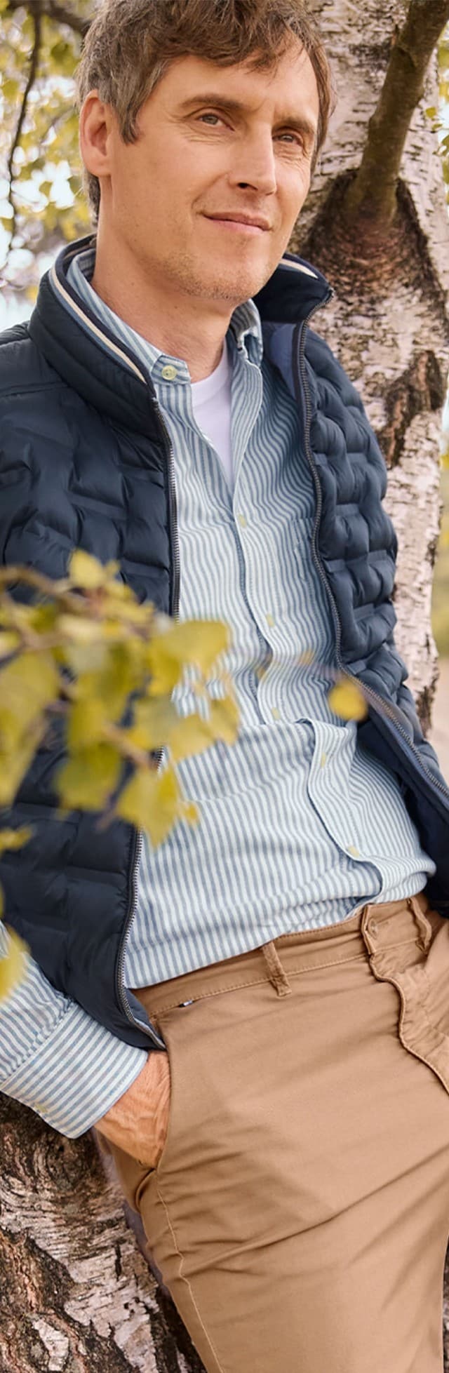 Eine Person trägt eine gestreifte Hemdbluse mit weißem T-Shirt darunter, dazu eine dunkelblaue Steppweste. Kombiniert mit beigefarbenen Hosen wirkt der Look sportlich und entspannt.