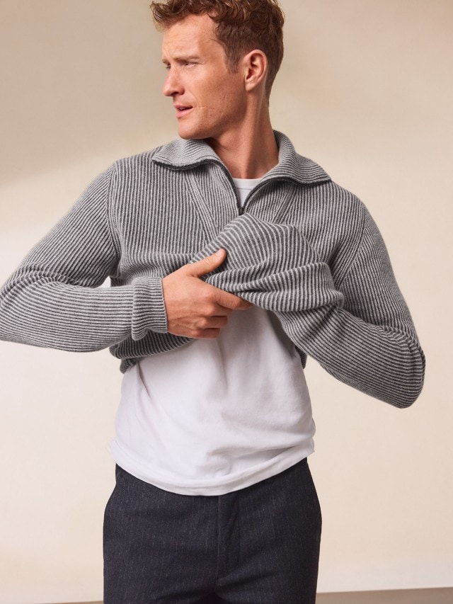 Ein Mann trägt einen grau gestreiften Strickpullover mit Reißverschlusskragen über einem weißen T-Shirt. Dazu kombiniert er eine dunkle Hose für ein sportlich-lässiges Outfit.