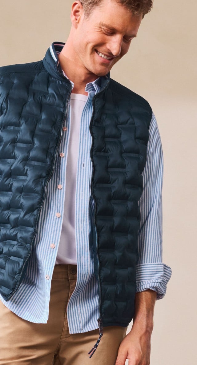 Ein Mann trägt eine gestreifte Hemdbluse mit weißem T-Shirt darunter und eine dunkelblaue Steppweste. Dazu kombiniert er eine beige Hose für ein sportlich-schickes Outfit.