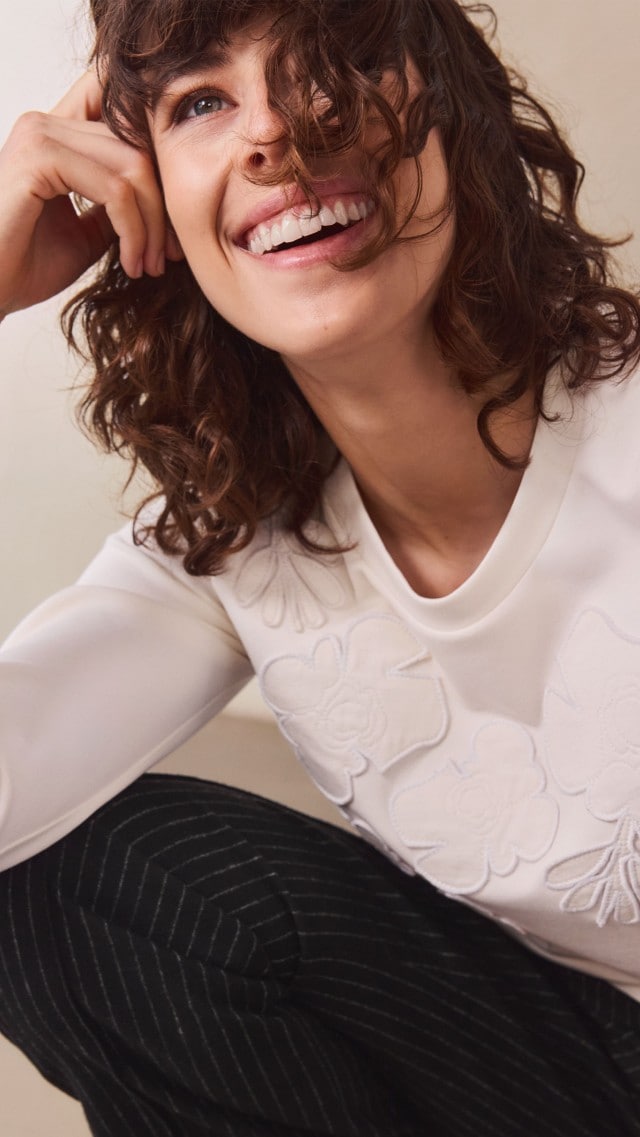 Eine Frau trägt einen cremefarbenen Pullover mit floralem Reliefmuster. Dazu kombiniert sie eine schwarze Hose mit dünnen weißen Streifen für einen modernen, eleganten Look.