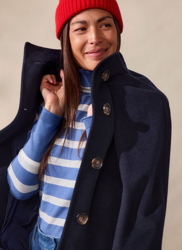 Eine Frau trägt ein blau-weiß gestreiftes Oberteil mit Rollkragen, kombiniert mit Jeans und einem dunkelblauen Mantel. Der Look ist leger, maritim inspiriert und modern.